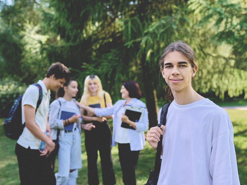 Male student 16, 17 years old with backpack, group of teenagers talking with teacher in school park. Back to school, back to college, high school, education, adolescence, teenage students concept